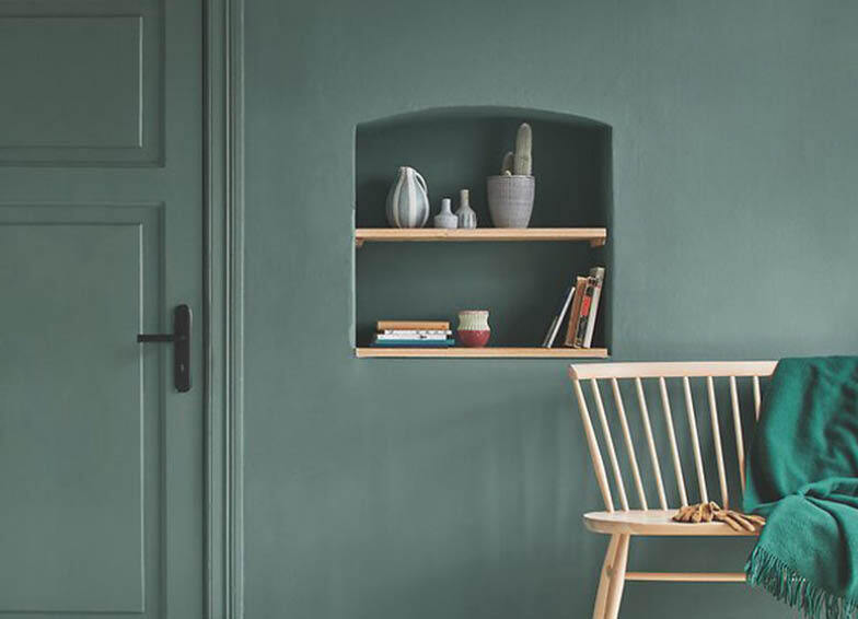 Farbmischservice: Stilvoll eingerichteter Raum mit einer Wand in einem gedämpften Grünton, einem Regal mit dekorativen Gegenständen und Büchern sowie einem hellen Holzstuhl mit einer grünen Decke.